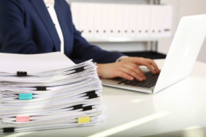 Stack_of_documents_and_woman_working_with_laptop_at_table_in_office,_closeup