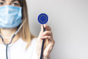 Young_woman_in_a_medical_mask_holds_a_stethoscope_with_the_flag_of_European_Union_on_a_light_background._Concept_of_medicine,_virus,_epidemic,_vaccination.