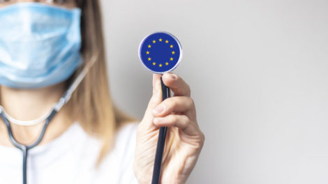 Young_woman_in_a_medical_mask_holds_a_stethoscope_with_the_flag_of_European_Union_on_a_light_background._Concept_of_medicine,_virus,_epidemic,_vaccination.