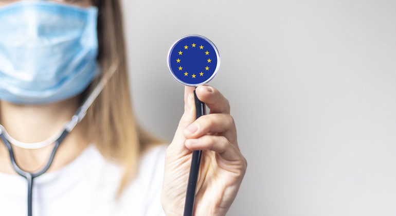 Young_woman_in_a_medical_mask_holds_a_stethoscope_with_the_flag_of_European_Union_on_a_light_background._Concept_of_medicine,_virus,_epidemic,_vaccination.