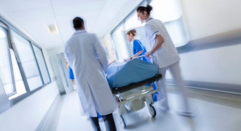 A_motion_blurred_photograph_of_a_patient_on_stretcher_or_gurney_being_pushed_at_speed_through_a_hospital_corridor_by_doctors_&_nurses_to_an_emergency_room