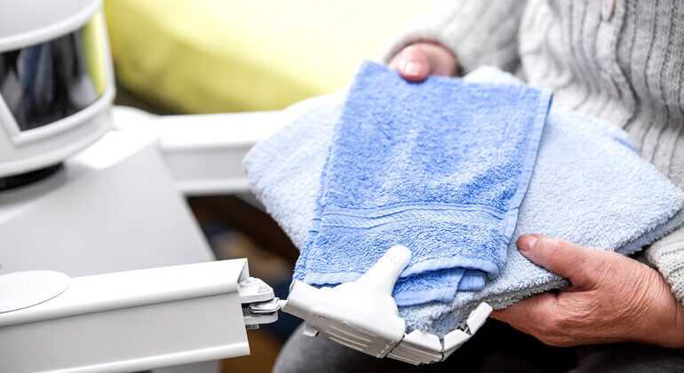 autonomous_caregiver_robot_is_holding_two_towels,_giving_them_to_an_senior_adult_woman_in_her_living_room,_concept_ambient_assisted_living